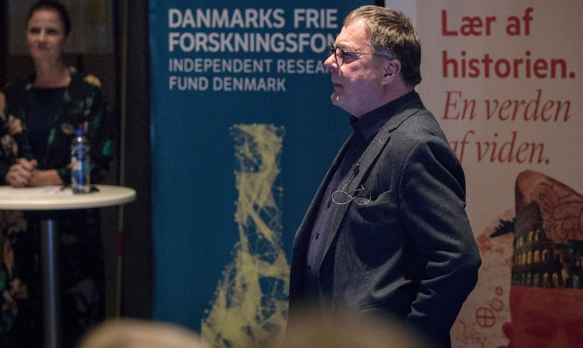 Foto af Peter Munk Christiansen foran en blå roll-up med Danmarks Frie Forskningsfonds logo og navn