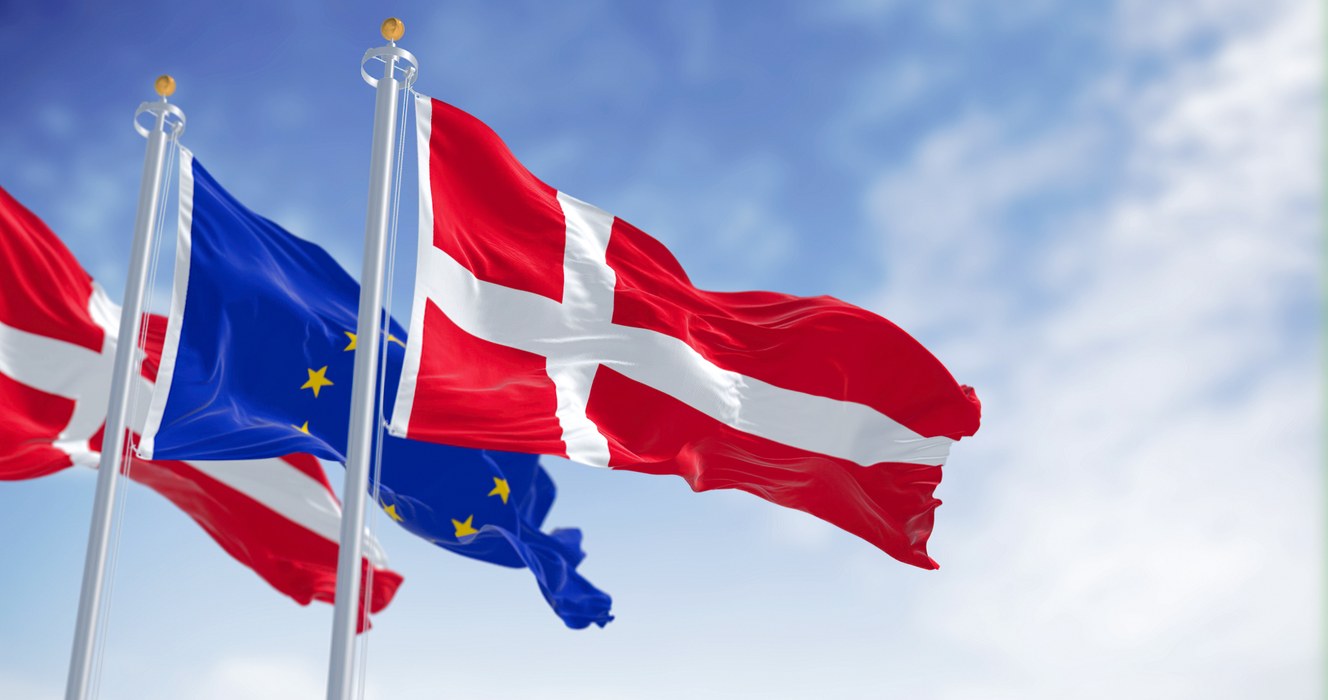 To danske flag med et europæiske flag mellem dem blafrer fra tre flagstænger på en lyseblå himmelbaggrund