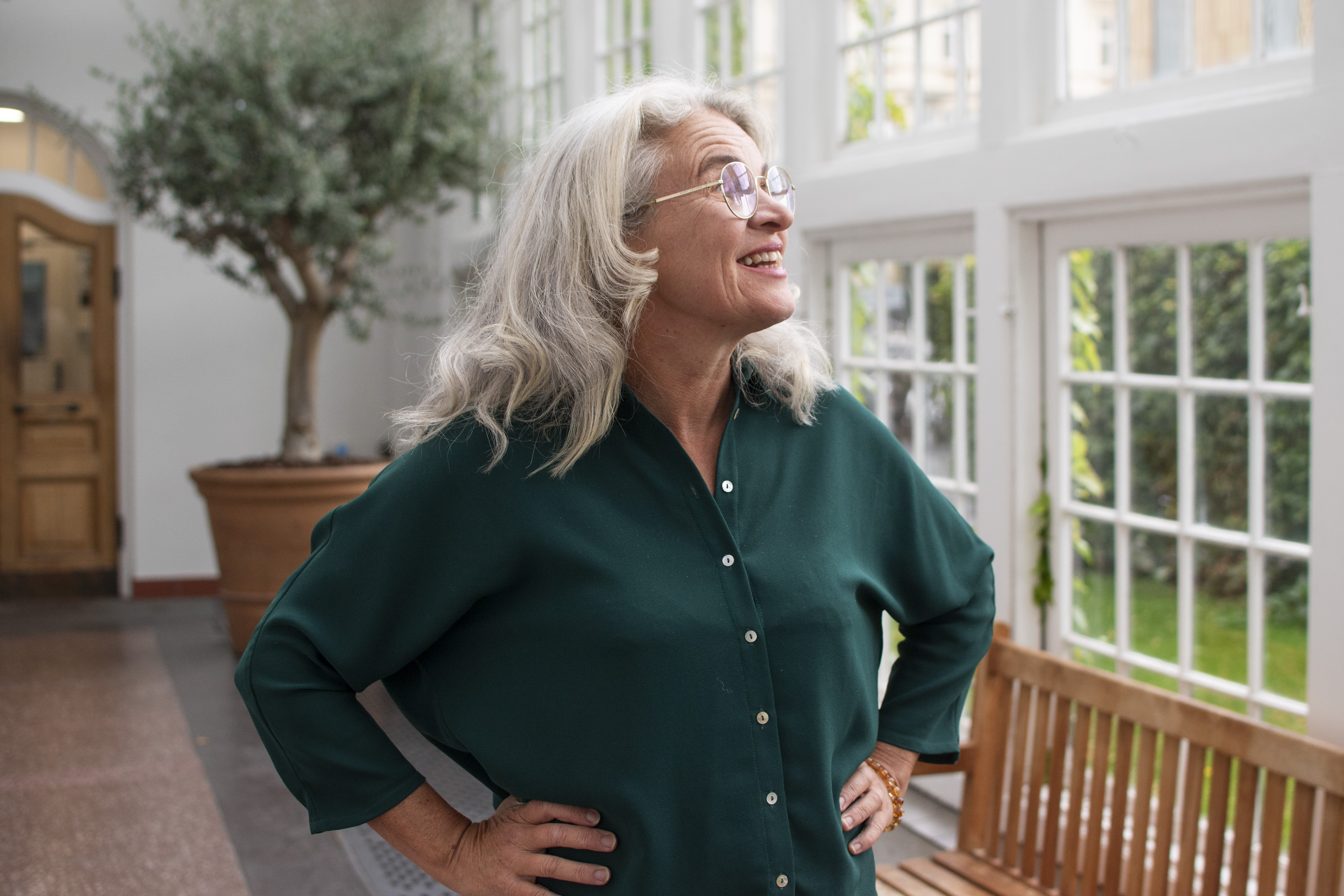 Foto af Lene Holm Pedersen der er i en mørkegrøn skjorte, med armene i siden og kigger til siden op på et vindue. Taget i hvad der ligner et orangeri med store vinduer og en stor grøn plante i baggrunden.