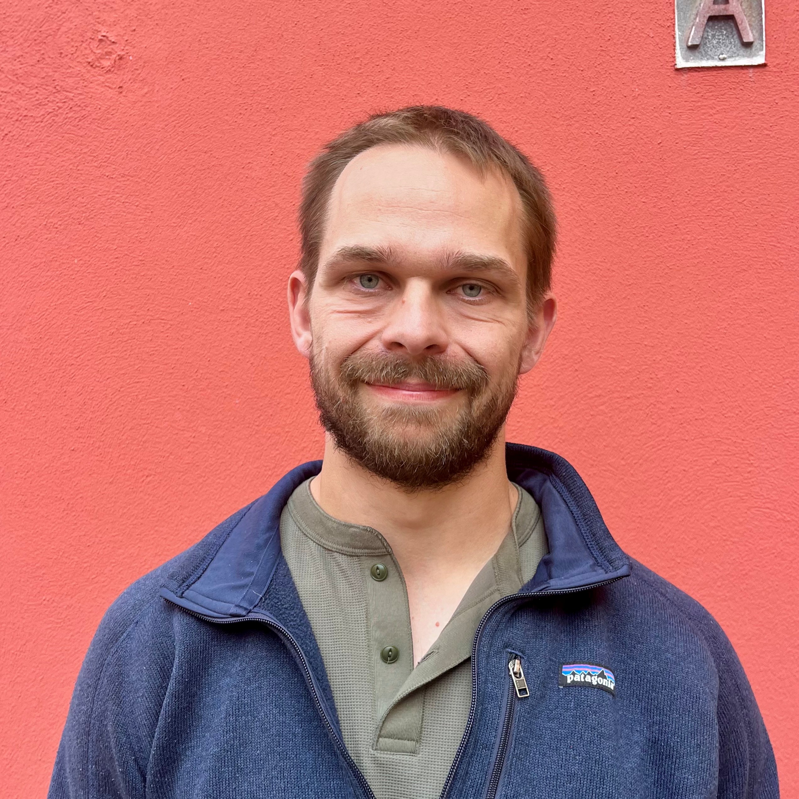 Foto af Thomas Lund Koch, der står op ad en rød væg og smiler let. 