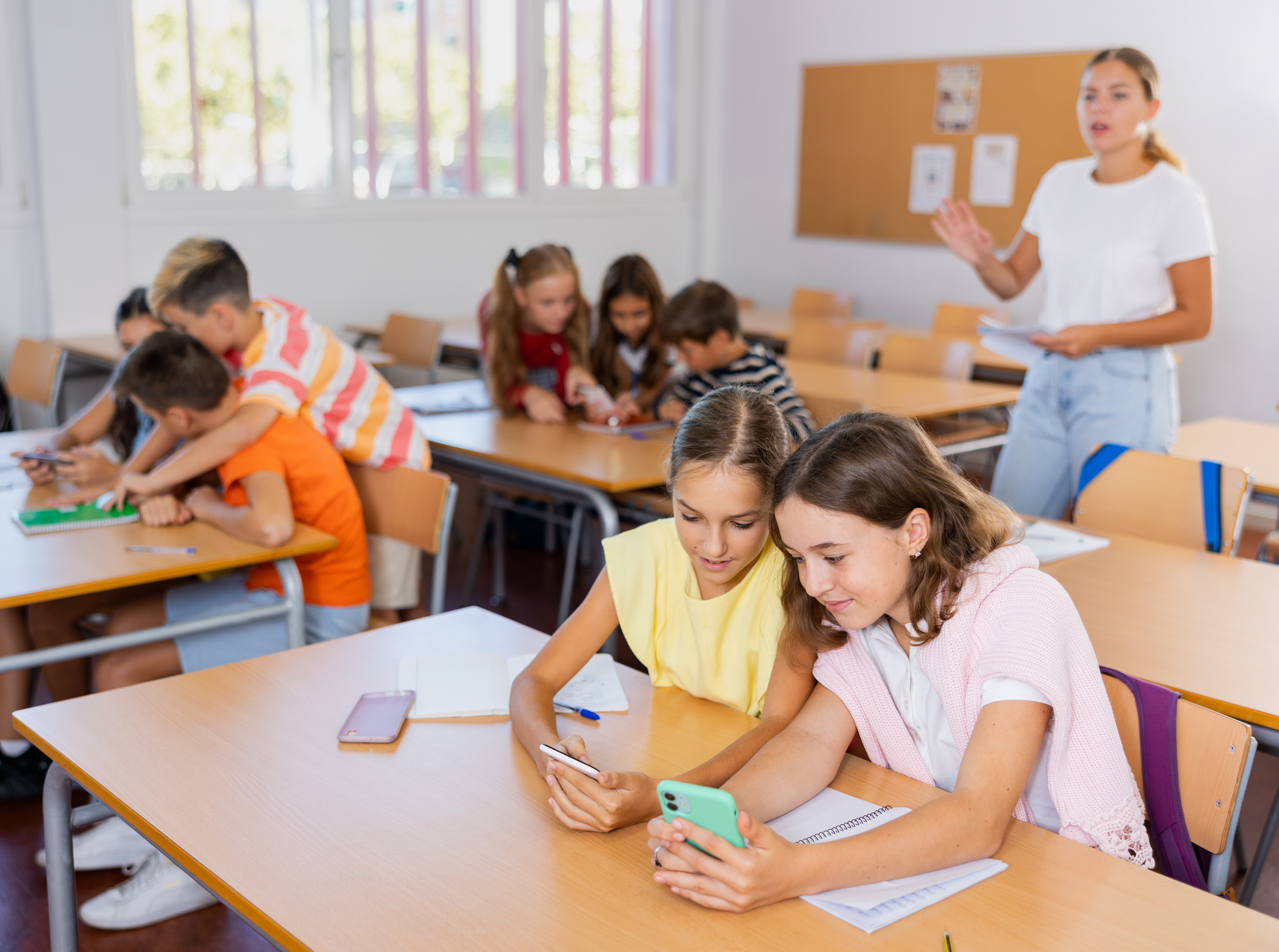 A classrom with pupils sitting in gruops working on a shared phone and a teacher i the bakground giving instructions