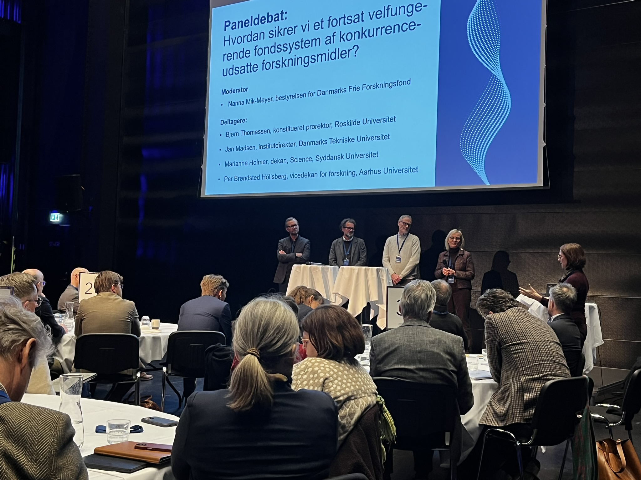 Fire paneldeltagere står på scenen under en pandeldebat ved dialogmøde 2026. I forgrunden af billedet ses andre deltagende ved dialogmødet, som sidder ved rundborde. Bag paneldeltagerne er et lærred.