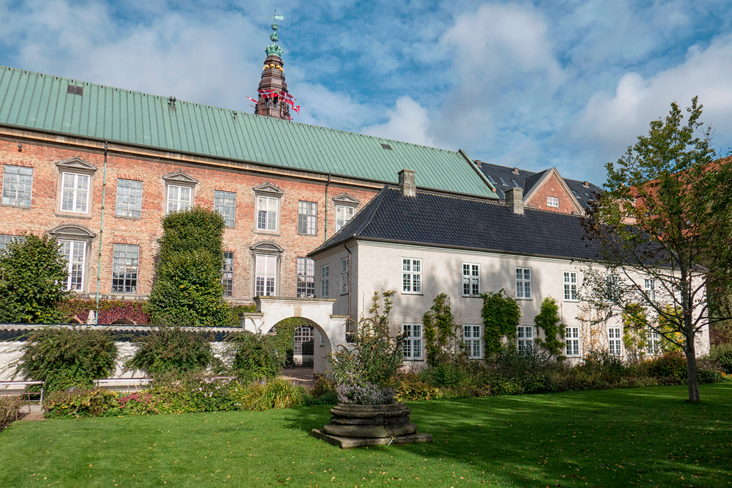 Foto af Rigsarkivet i København med sine røde, gamle murstensmure. Det er set fra haven i gården, så der er en græsplæne forrest i billedet og Rigsarkivets røde mure i solskin bagerst.