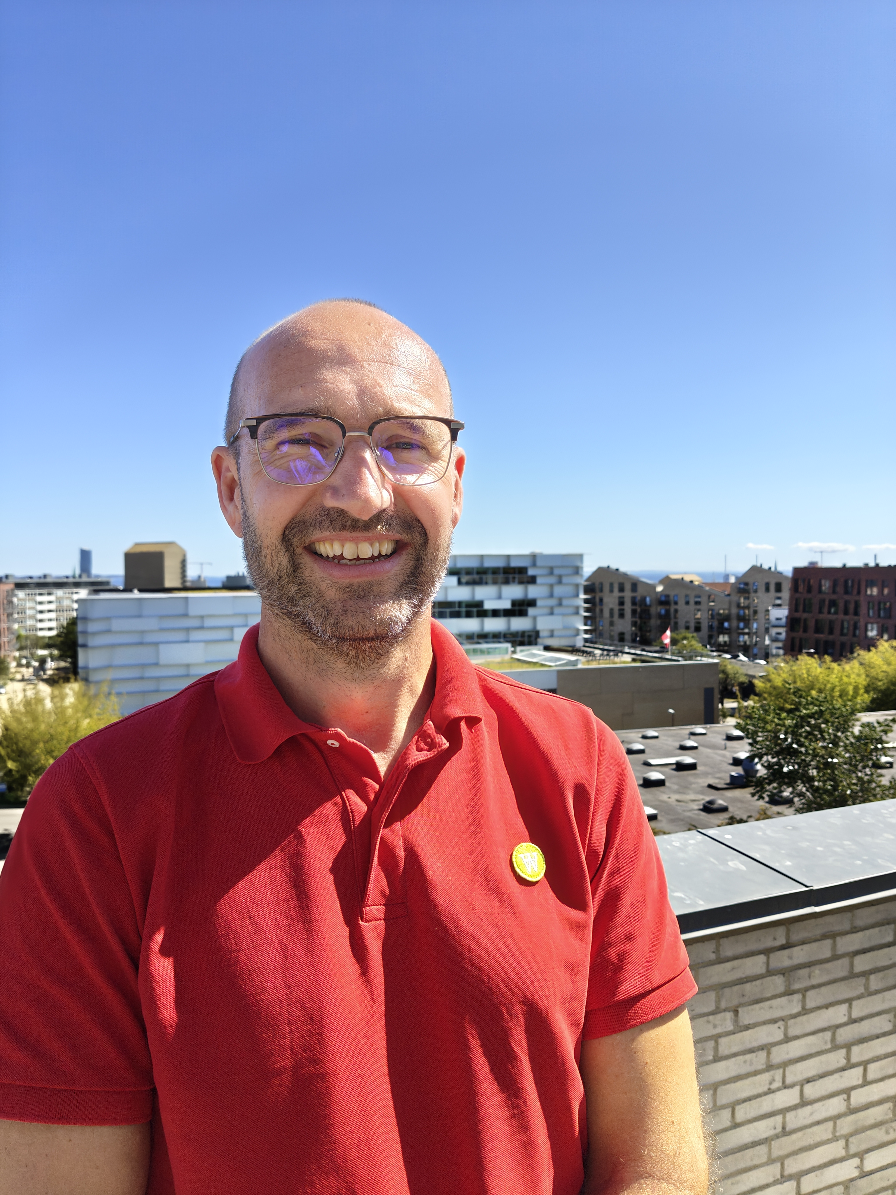 Foto af Jakob Juul Larsen taget udenfor med en storby i baggrunden. Solen skinner, og Jakob har en rød polo t-shirt på.